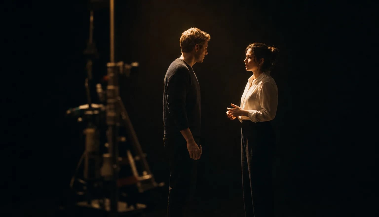 Two performers mid-conversation in a single pool of warm light on an otherwise dark soundstage